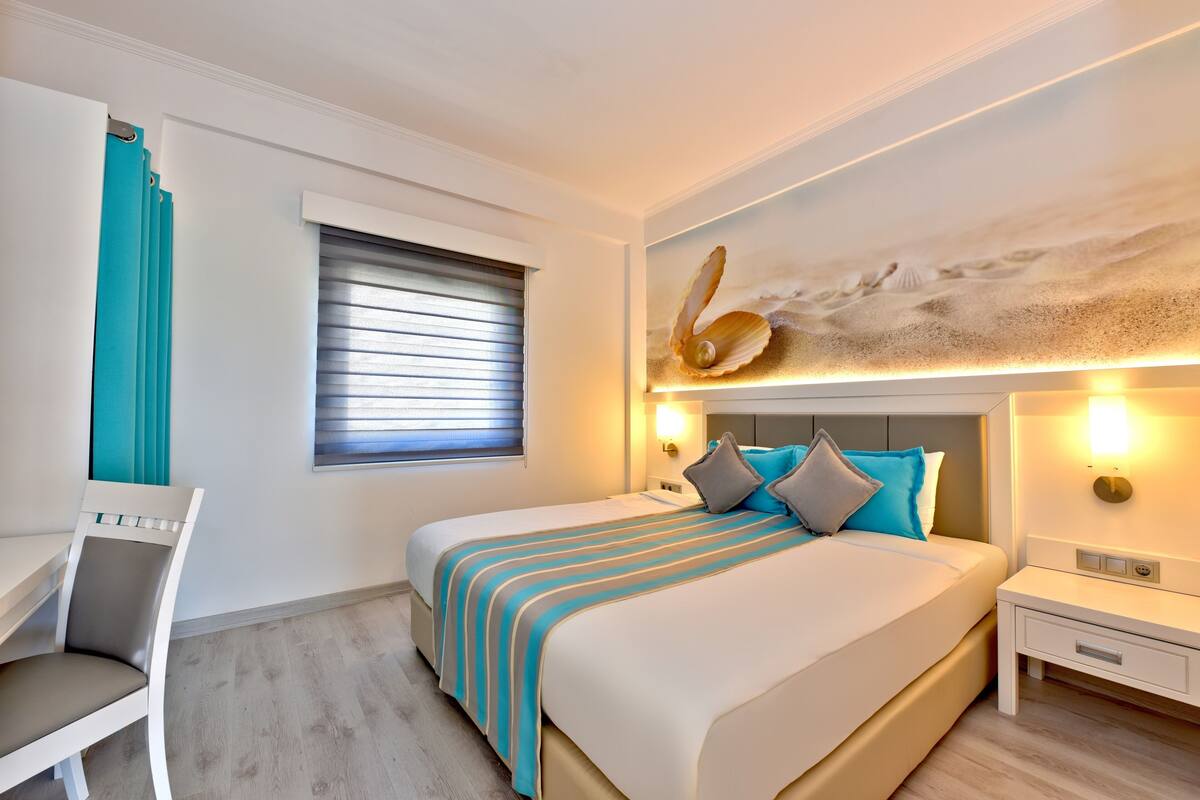 A modern hotel room with a large bed, a desk, and a window with blinds.