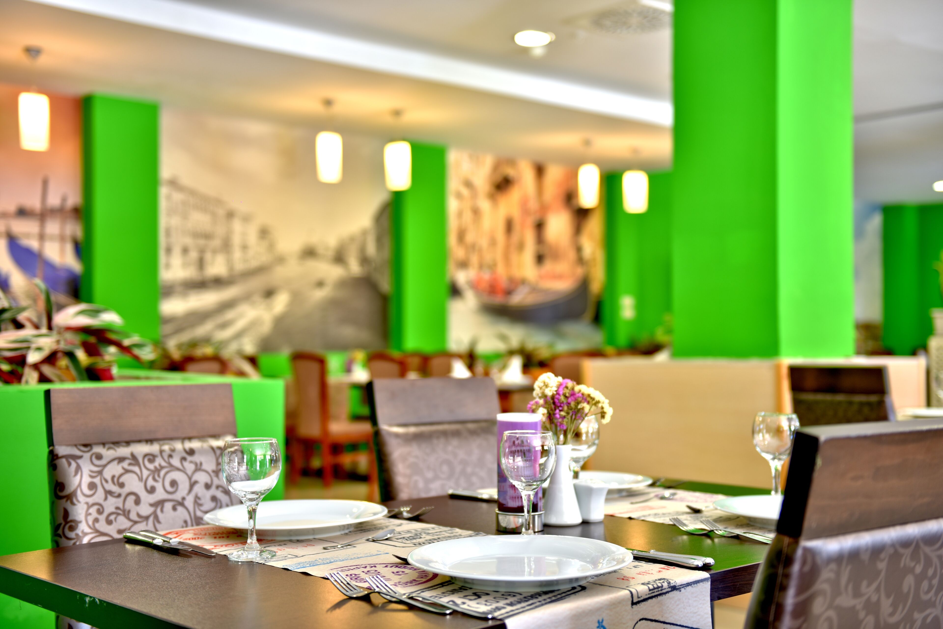 Un comedor con paredes verdes, un mural y mesas ordenadas con platos, vasos y cubiertos.