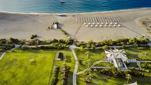 Vista aérea de un complejo turístico costero con playa, zona de estacionamiento y un edificio central con un diseño de techo singular.