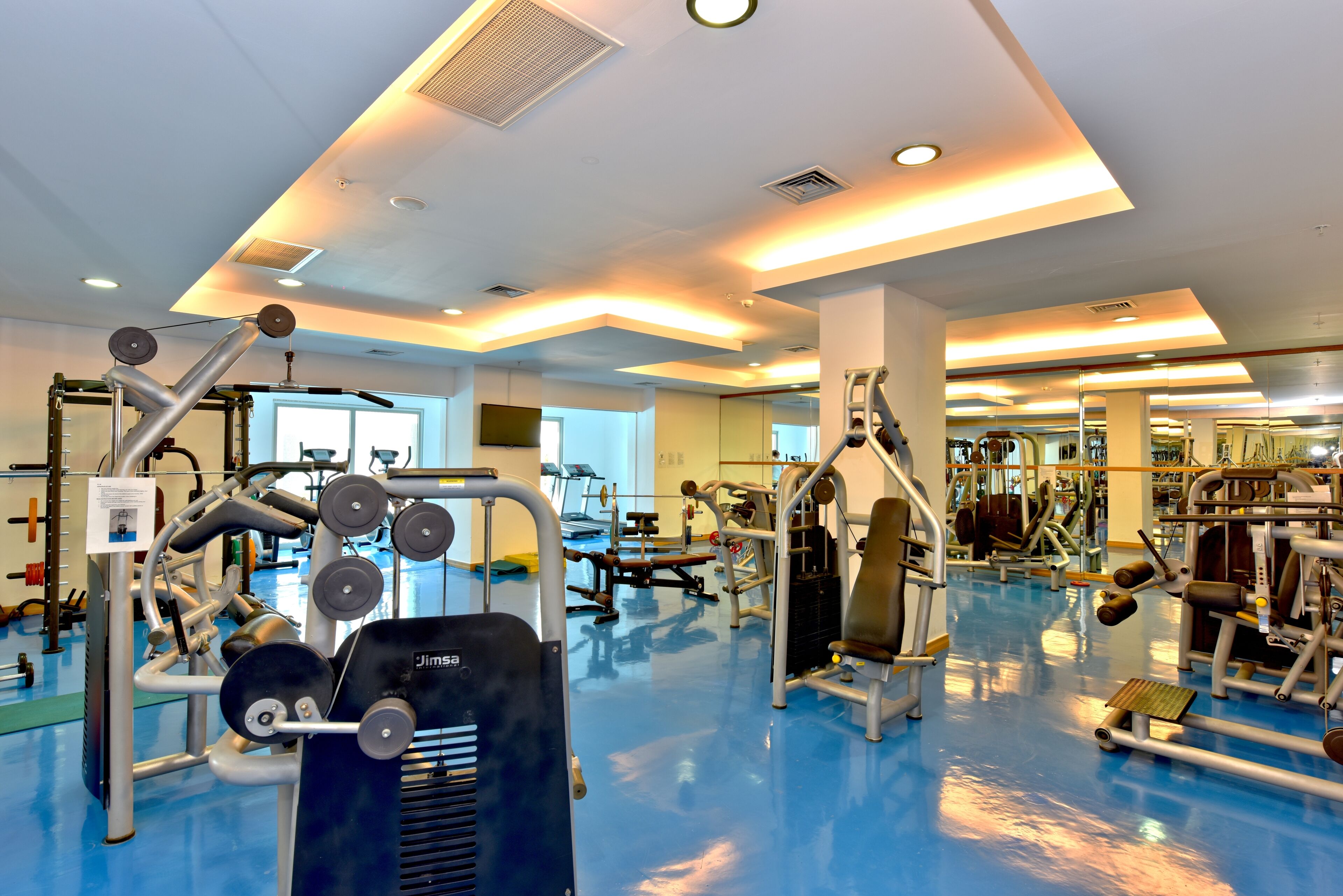 Ein Fitnessstudio mit unterschiedlichen Trainingsgeräten, einem blauen Boden und einer Decke mit eingelassenen Leuchten.