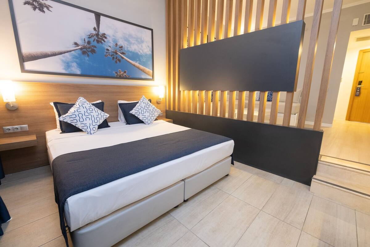 A modern hotel room with a large bed, a headboard with vertical slats, a wall-mounted TV, and a framed picture of palm trees.