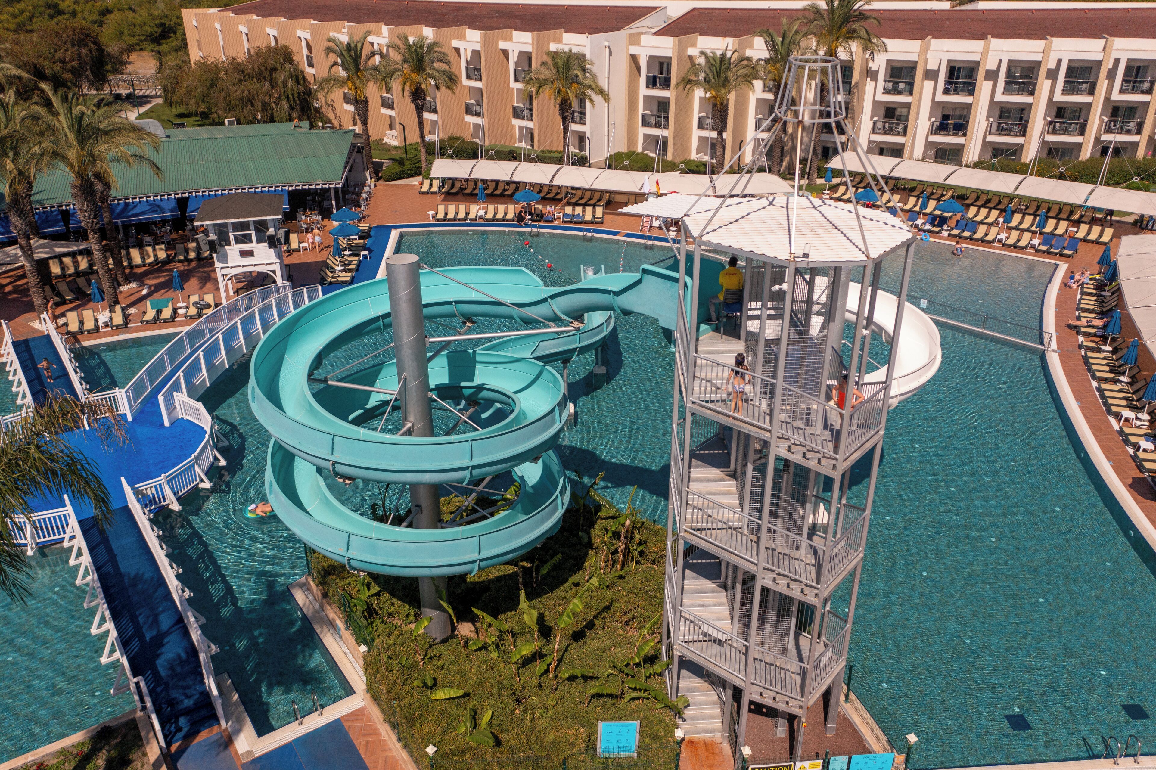A water park with multiple slides and a pool area, surrounded by a hotel with balconies and lounge chairs.