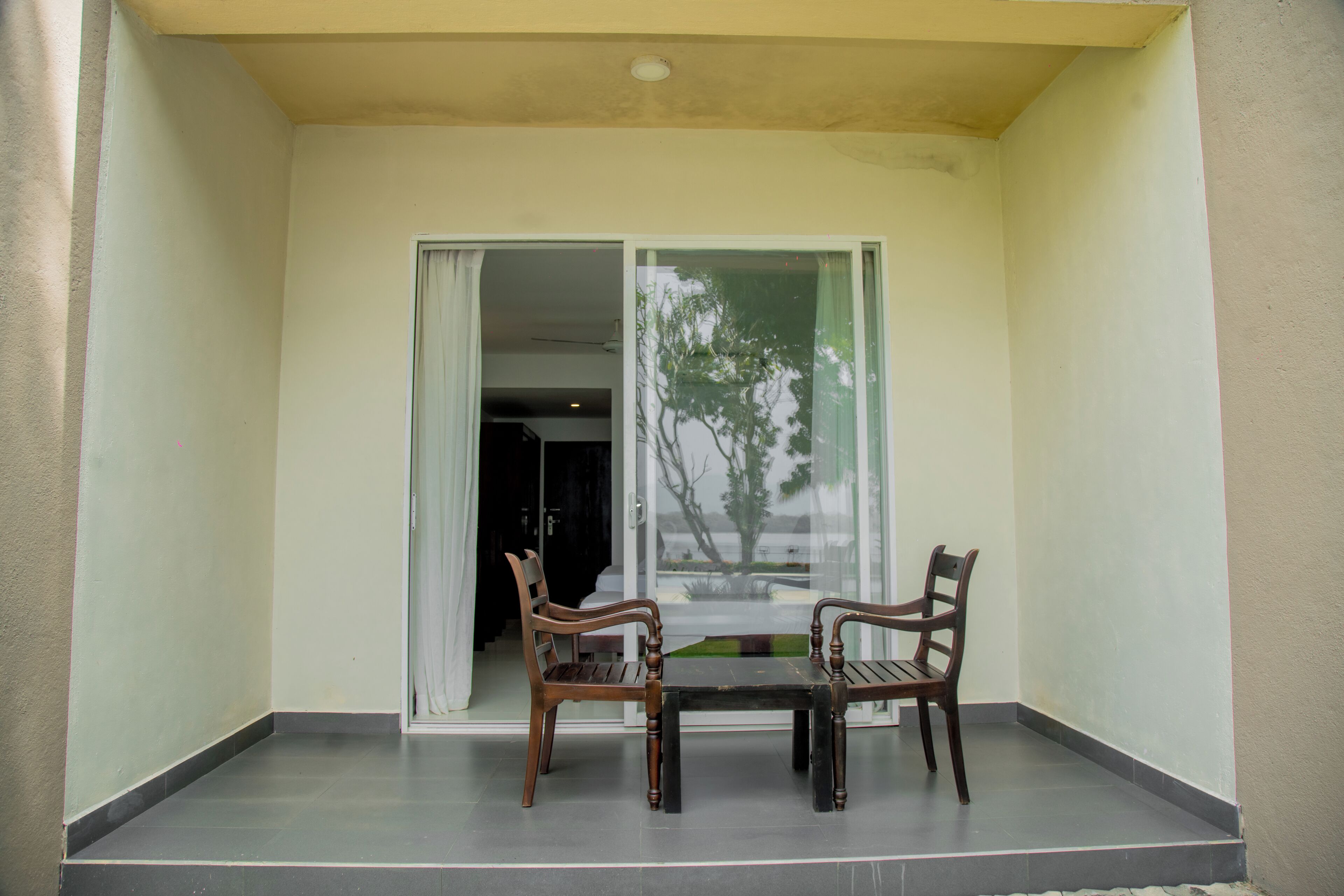 A balcony with two wooden chairs and a table, a sliding glass door, and a view of a tree and building.