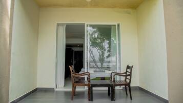 A balcony with two wooden chairs and a table, a sliding glass door, and a view of a tree and building.