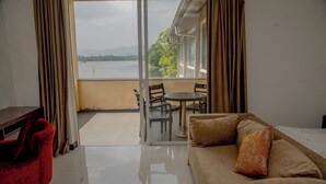 A room with a sofa, a table, and a chair, a balcony with a view of a lake and mountains, and a glass door. - Lavendish Wild Safari Resort (Maraka)