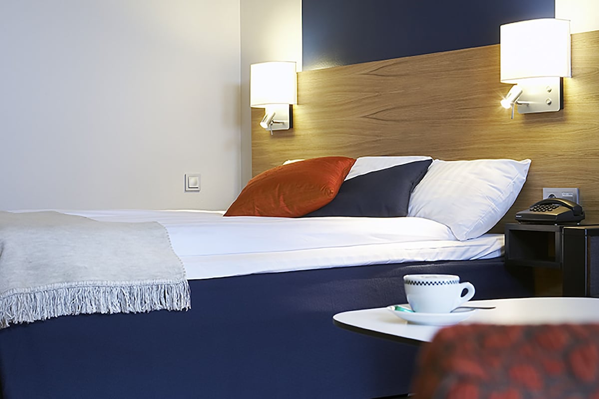 Una habitación de hotel con cama, mesita de noche, lámpara y una taza sobre la mesa.