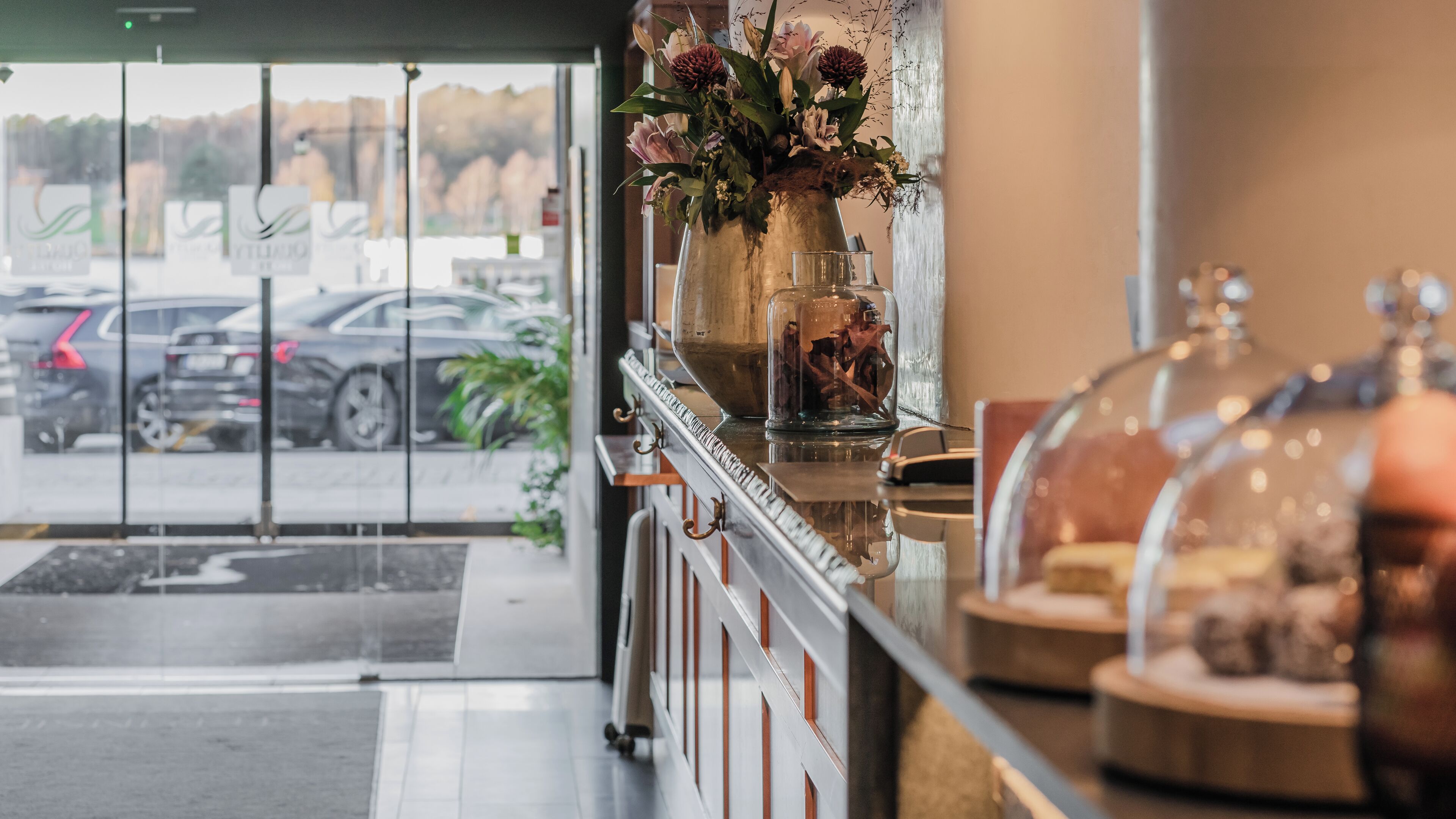 A hotel reception area with a glass door leading outside, a vase with flowers on the counter, and a display case with items.