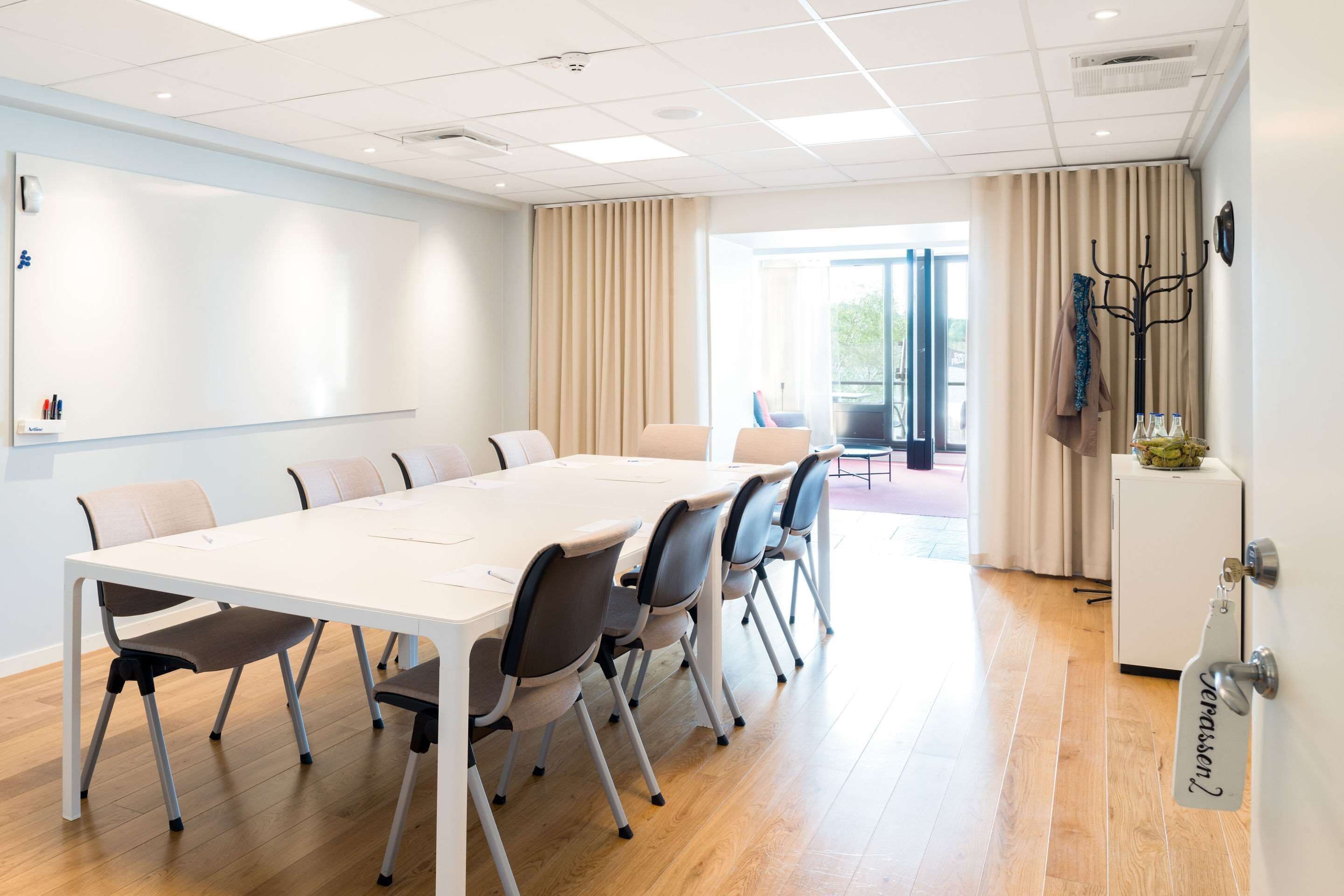 A conference room with a white table, six chairs, a whiteboard, and a door with a coat rack.