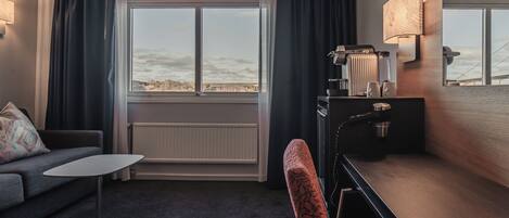 Habitación de hotel con sofá, una mesita, escritorio con lámpara y vistas al exterior.