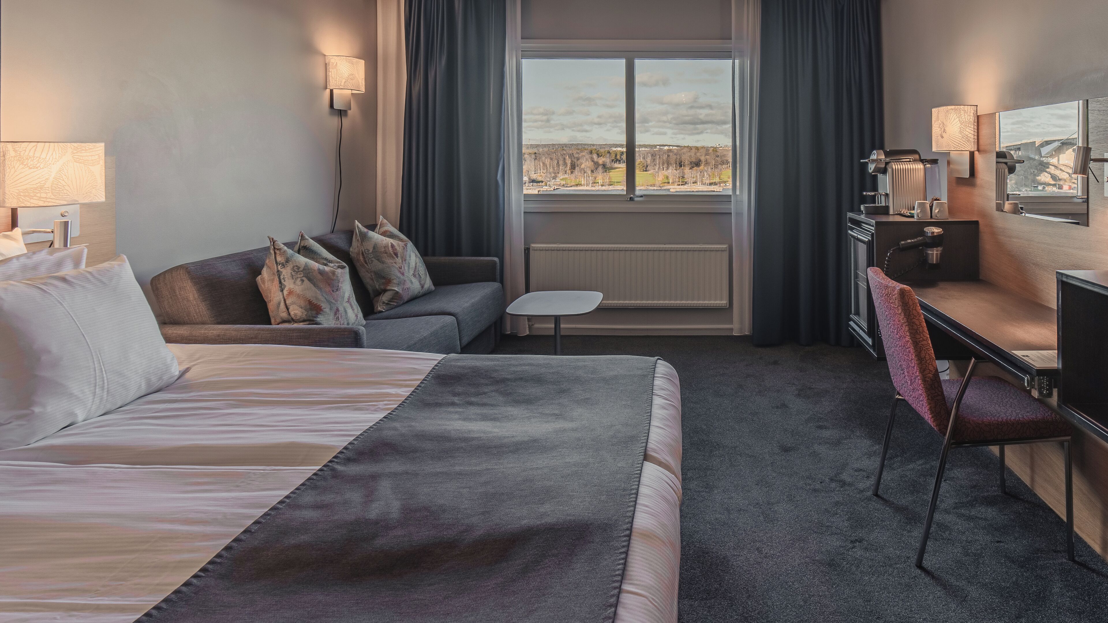 A hotel room with a bed, a desk, a chair, a small table, and a view of the outdoors.