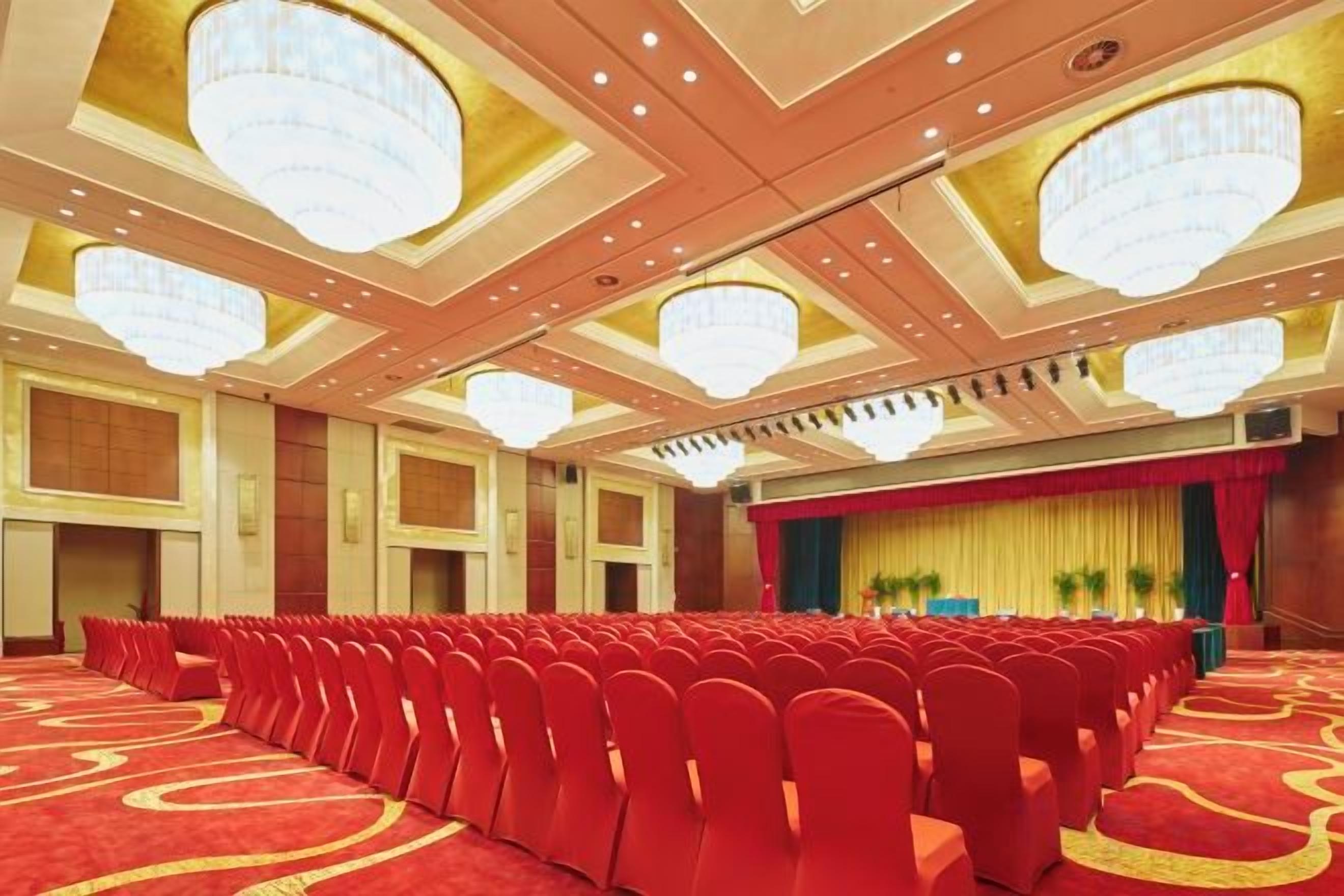 A large conference room with rows of chairs, a stage with a curtain, and chandeliers.