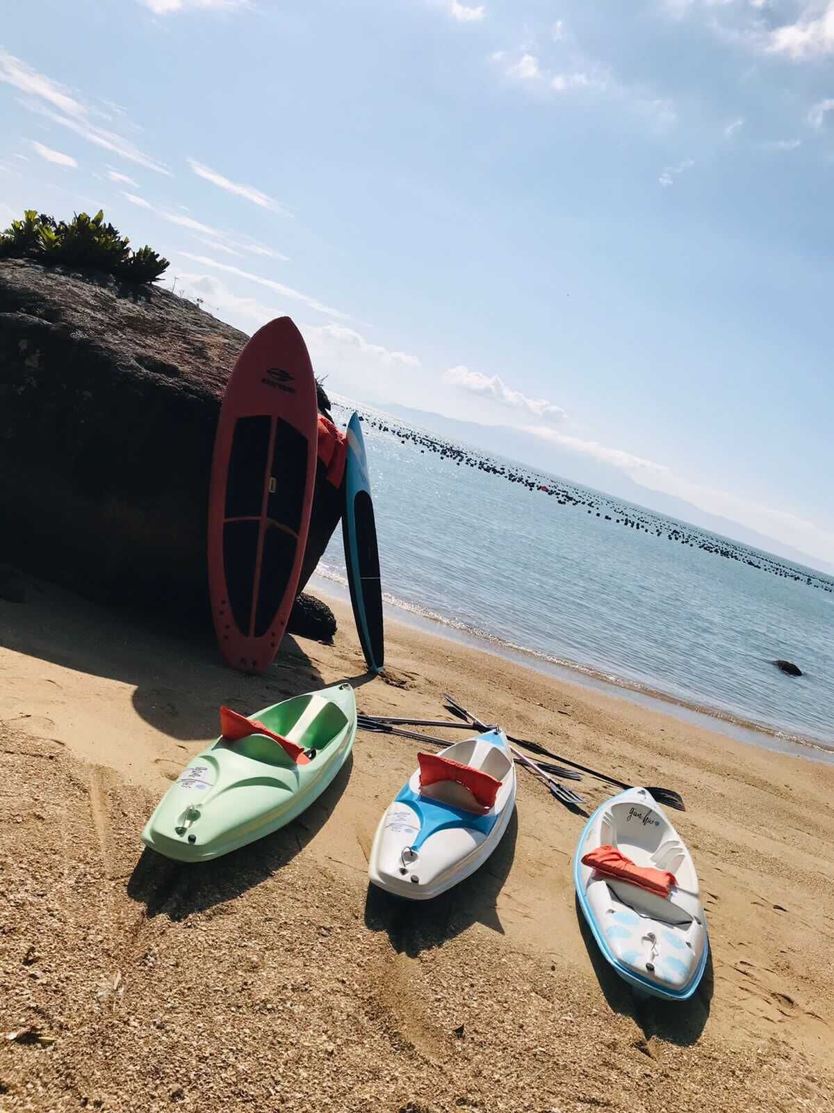 Kajaks en een surfplank op een zandstrand, met een rotsachtige klif en rustig water op de achtergrond.