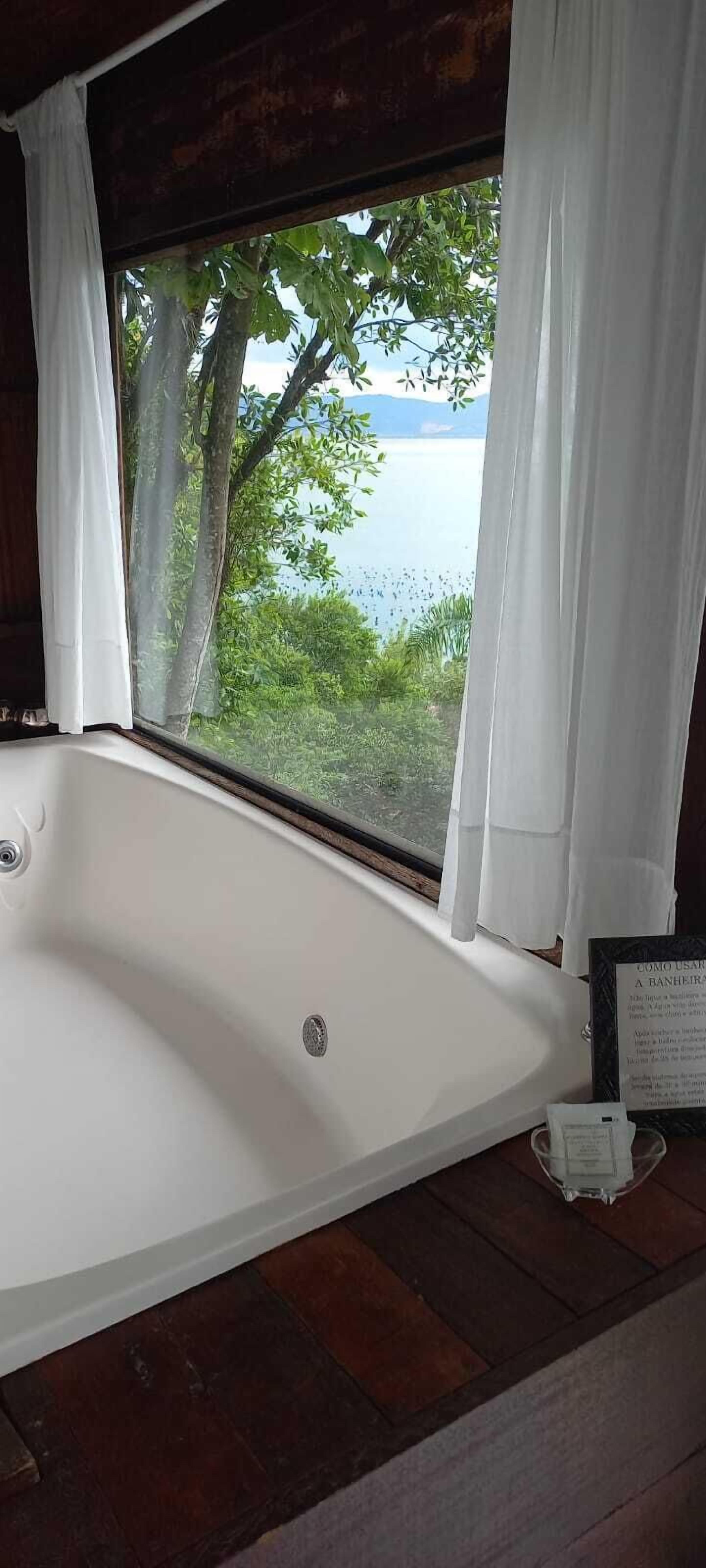 A large, empty bathtub with a view of a lake and trees through a window with white curtains.