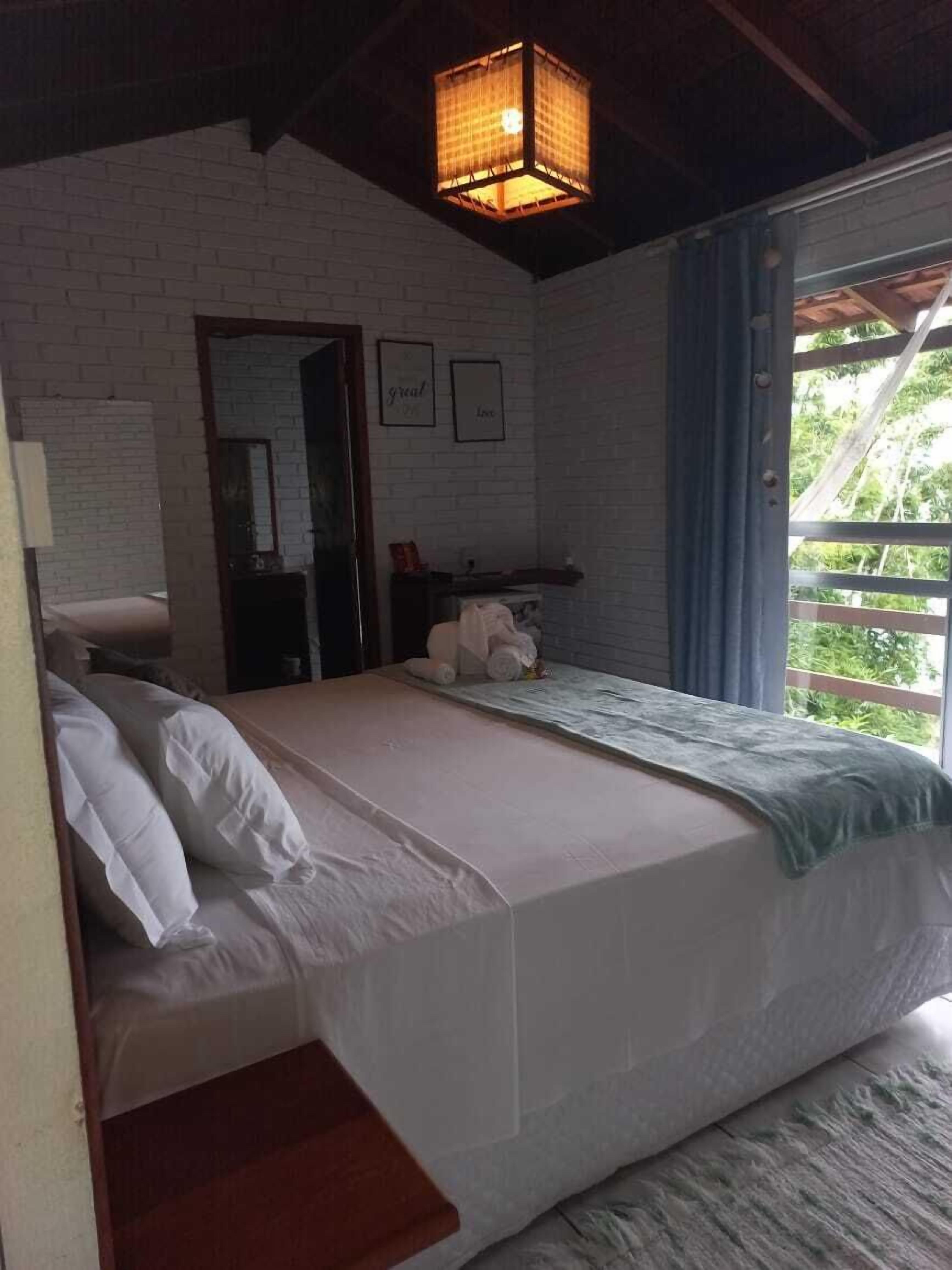 A bedroom with a bed, a nightstand, a mirror, a window with curtains, and a hanging light fixture.