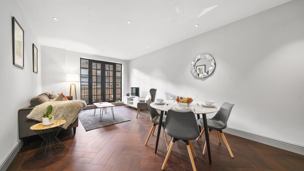 A modern living room with a dining area, wooden flooring, and a large window with blinds.