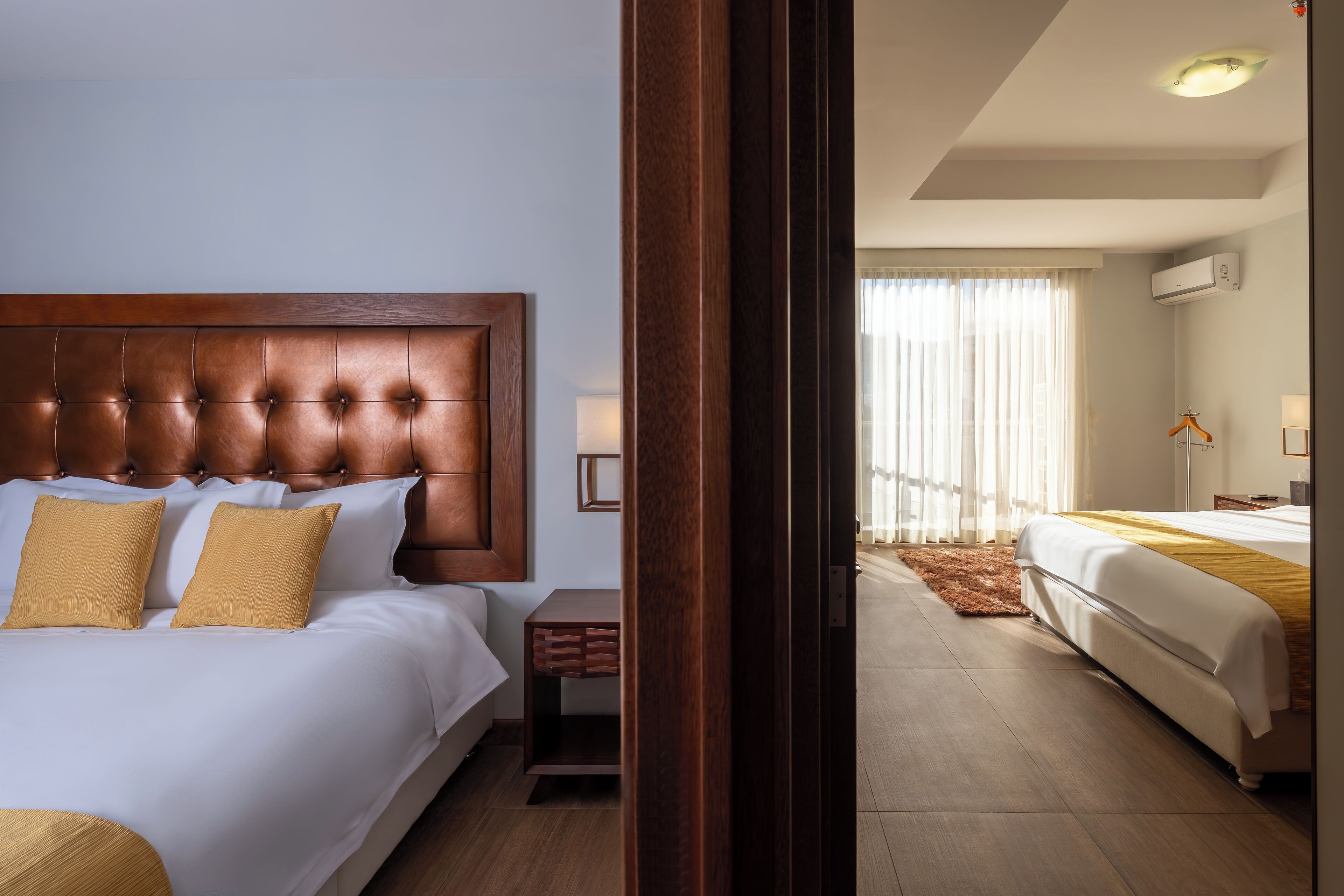 Quarto de hotel com uma cama grande, uma cama menor, uma mesinha de cabeceira de madeira e vista para outro quarto através de uma porta aberta.