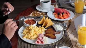 Un desayuno con huevos revueltos, jamón, queso y tostadas, acompañado de una taza de café y un vaso de zumo de naranja.