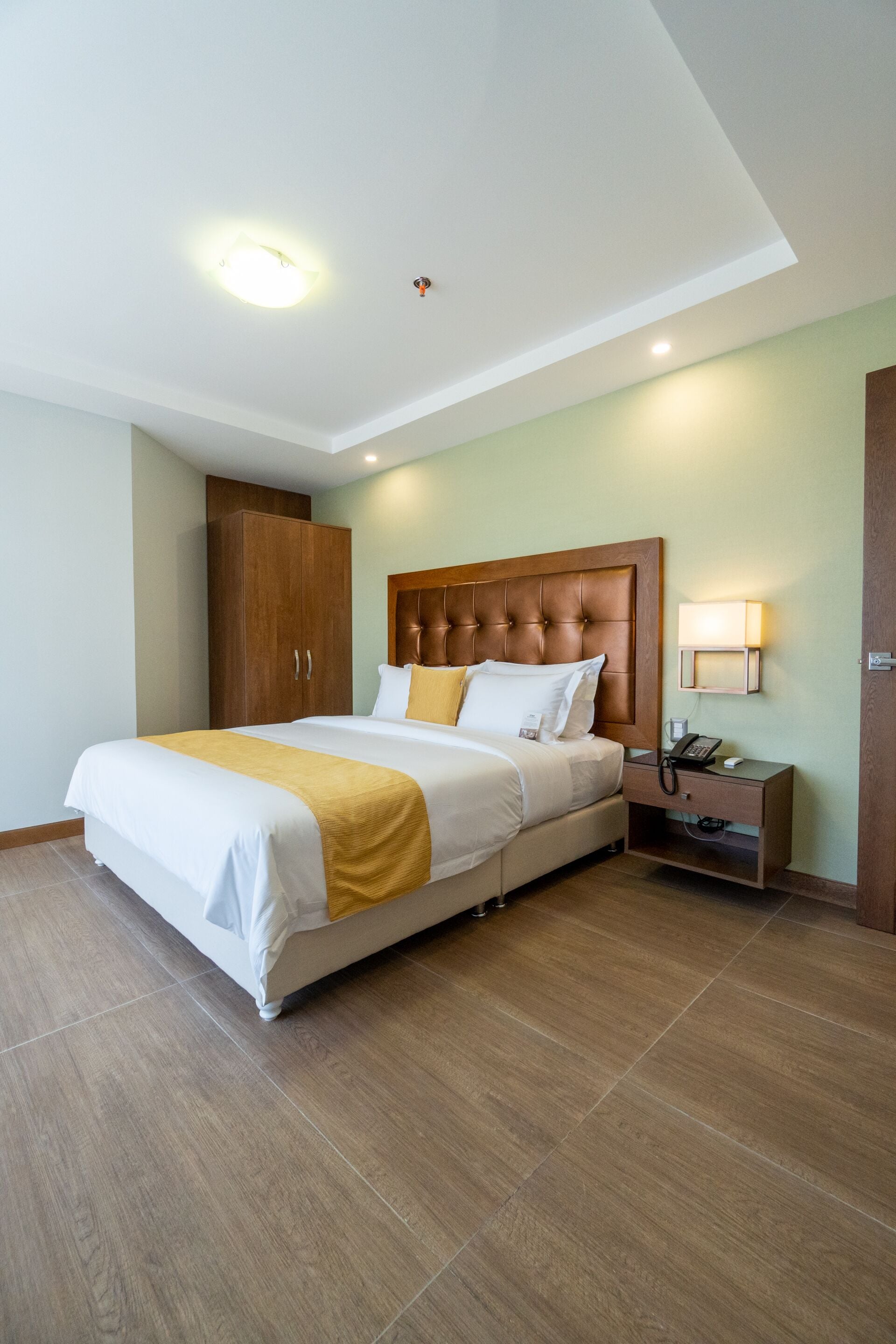 Una habitación de hotel con una cama grande, cabecera de madera, mesita de noche con lámpara y un armario.