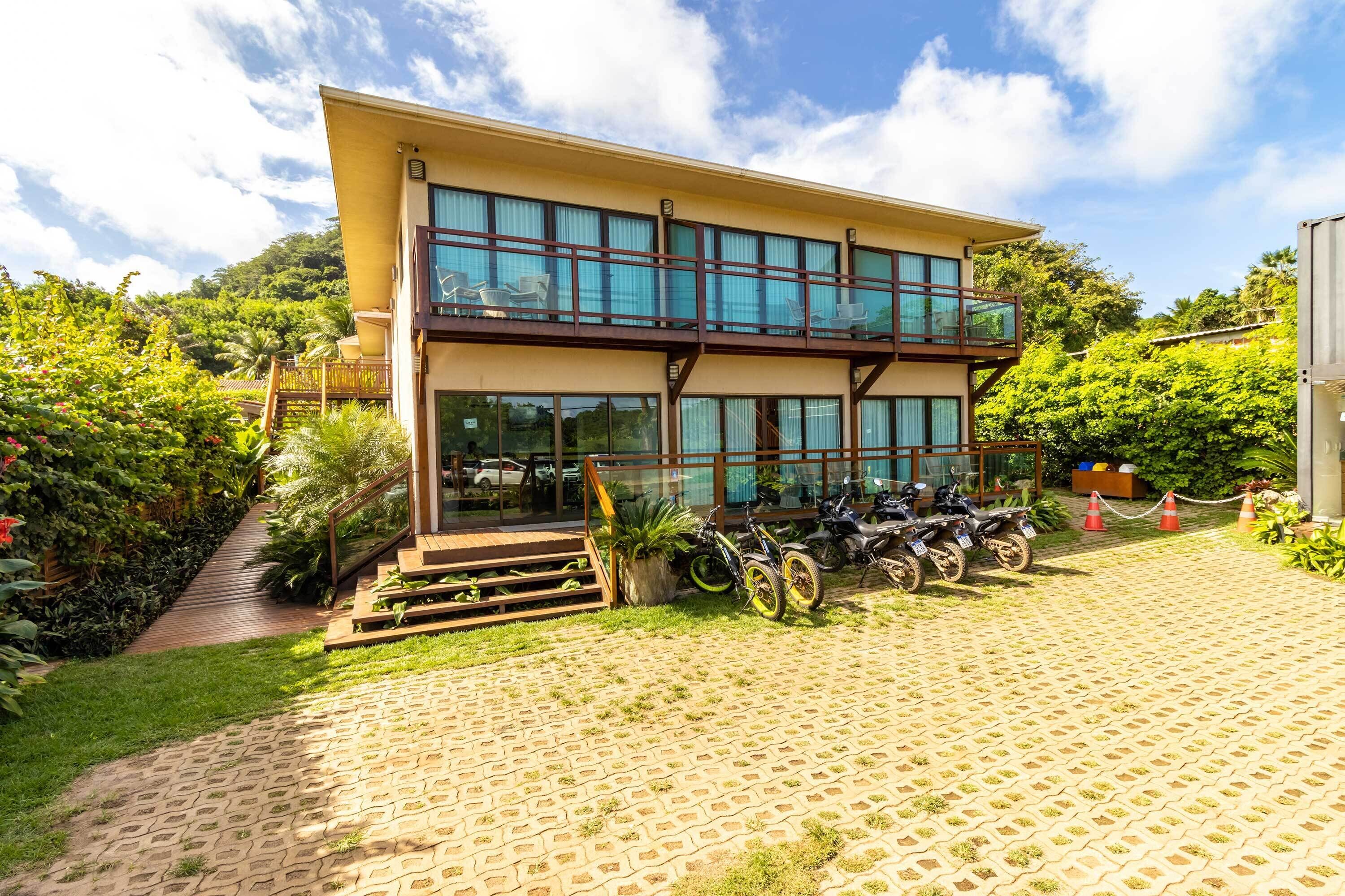 A two-story building with a balcony, surrounded by lush greenery and a paved driveway.