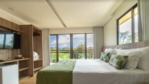 A modern bedroom with a large bed, a TV mounted on a wooden cabinet, and a balcony with a view of greenery. - Pousada Morro do Farol (Fernando de Noronha)