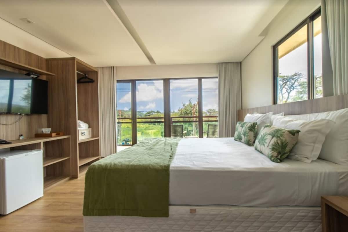 Ein modernes Schlafzimmer mit einem großen Bett, einem an einer Holzeinrichtung montierten Fernseher und einem Balkon mit Blick auf Grünflächen.