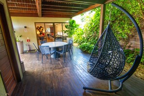 An outdoor patio with a round table and chairs, surrounded by lush greenery and a hanging chair.