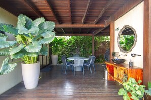 An outdoor patio with a round table and chairs, surrounded by large potted plants and a wooden ceiling.