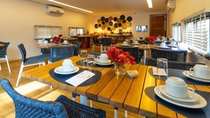 A modern dining area with wooden tables and blue chairs, a wall-mounted decorative item, and a window with blinds. - Pousada Morro do Farol (Fernando de Noronha)