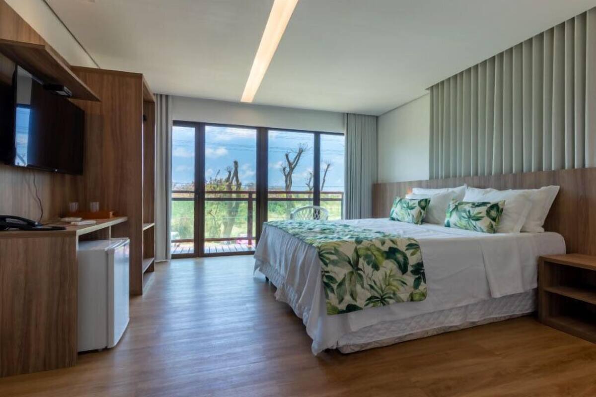 Ein modernes Hotelzimmer mit einem großen Bett, einem Schreibtisch, einem Fernseher und einem Balkon mit Blick auf Bäume.