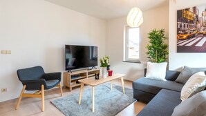 A modern living room with a flat-screen TV, a grey sofa, a wooden coffee table, and a large potted plant. - Jette Duplex Expo (Brussels)
