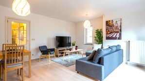 A modern living room with a wooden dining table, a grey sofa, a TV, and a large window with a cityscape view. - Jette Duplex Expo (Brussels)