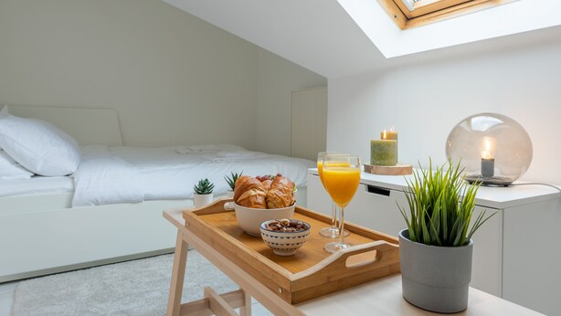 A modern bedroom with a skylight, a breakfast tray with croissants, a glass of orange juice, and a potted plant. - Jette Duplex Expo (Brussels)