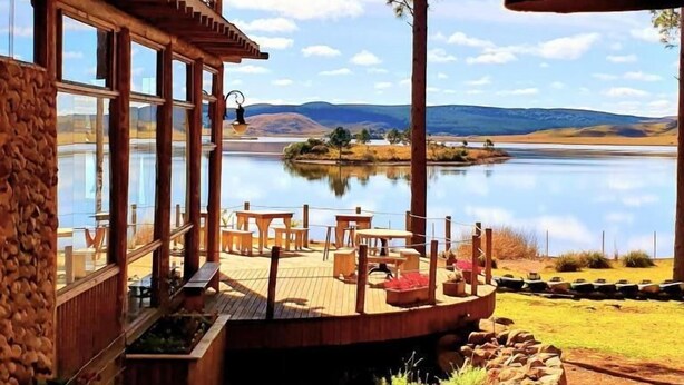 A wooden cabin with a deck overlooking a lake, featuring outdoor seating and a stone wall.