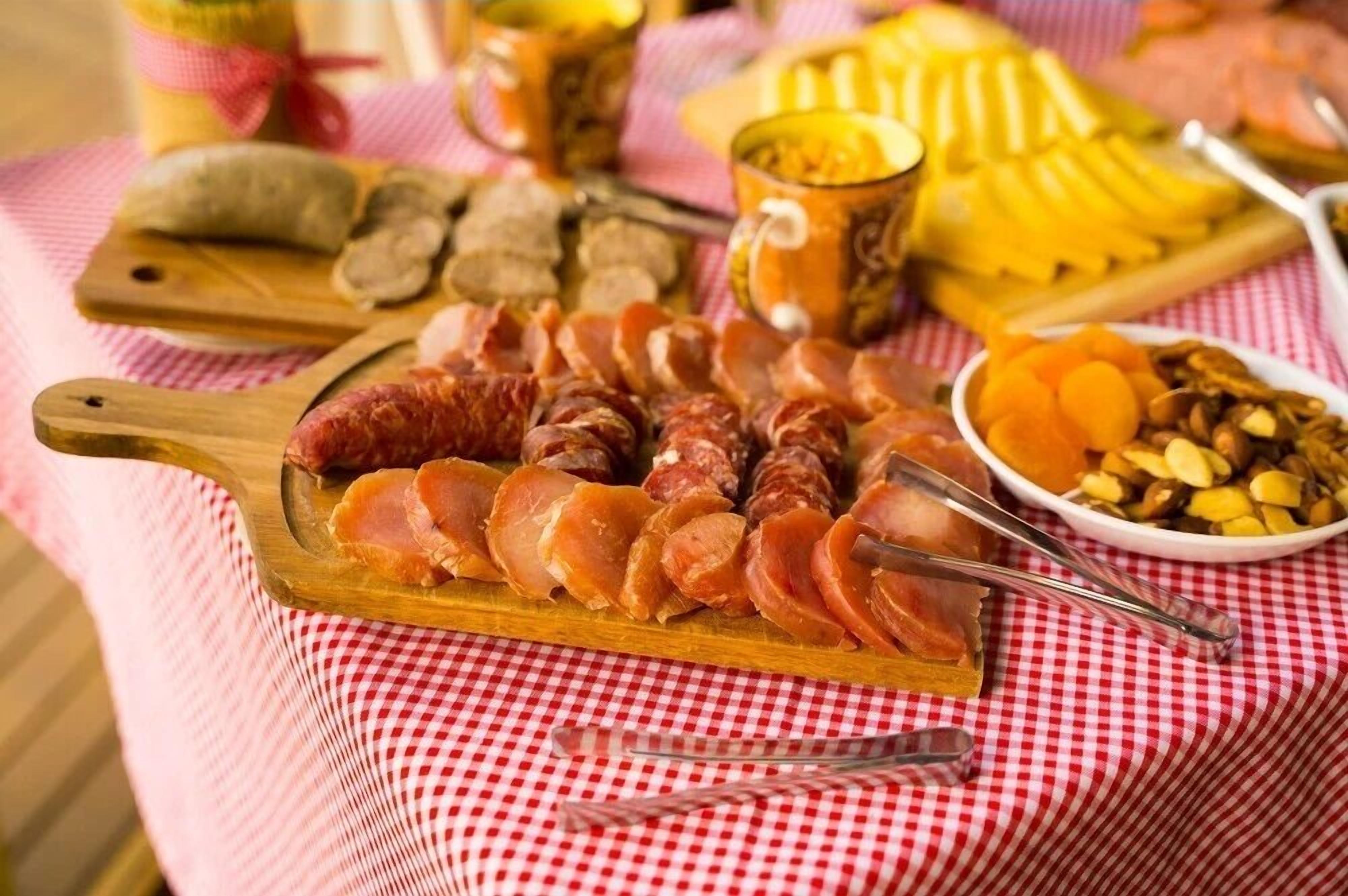 Una tabla de fiambres con diversos embutidos y quesos sobre una tabla de madera, acompañada de un cuenco con frutos secos y frutas deshidratadas sobre un mantel a cuadros.