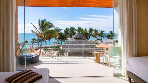 A room with a large glass window overlooking a beach with palm trees and a thatched-roof structure. - qema Mancora (Máncora)