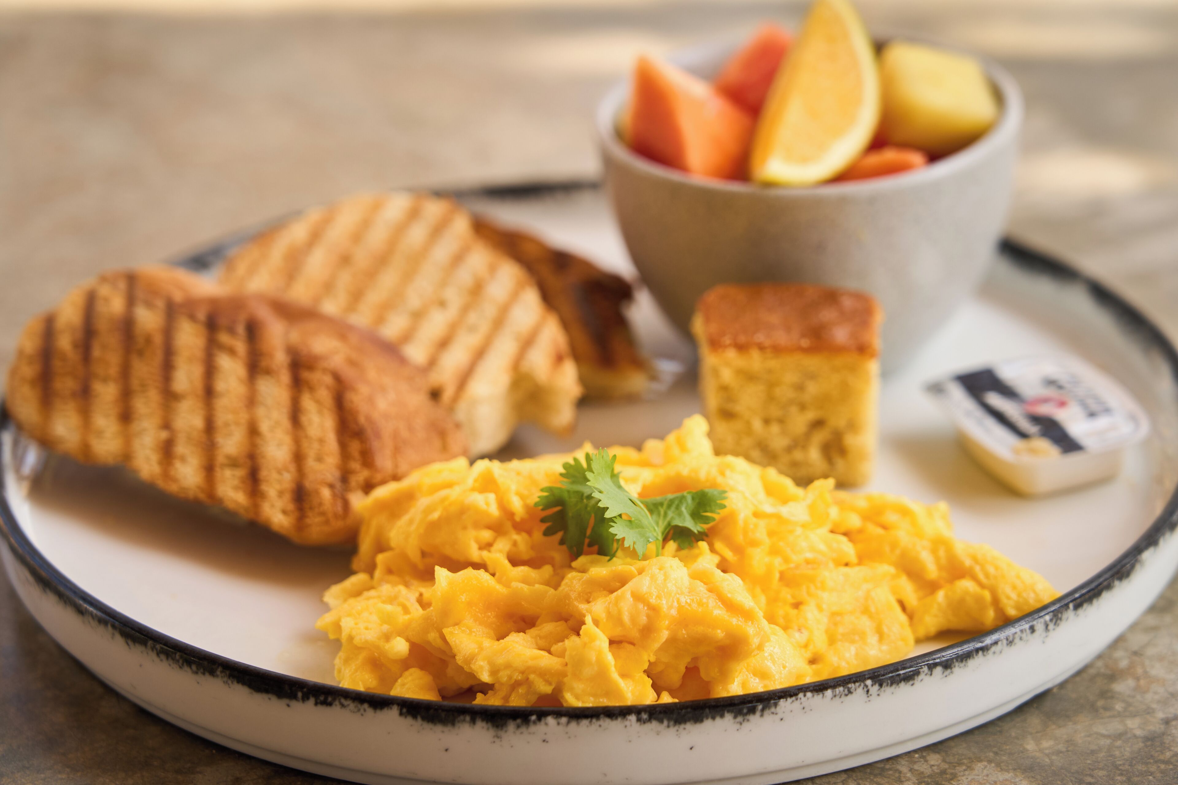 Un plato con huevos revueltos, un sándwich a la plancha, un trozo de pan de maíz y un bol de fruta.