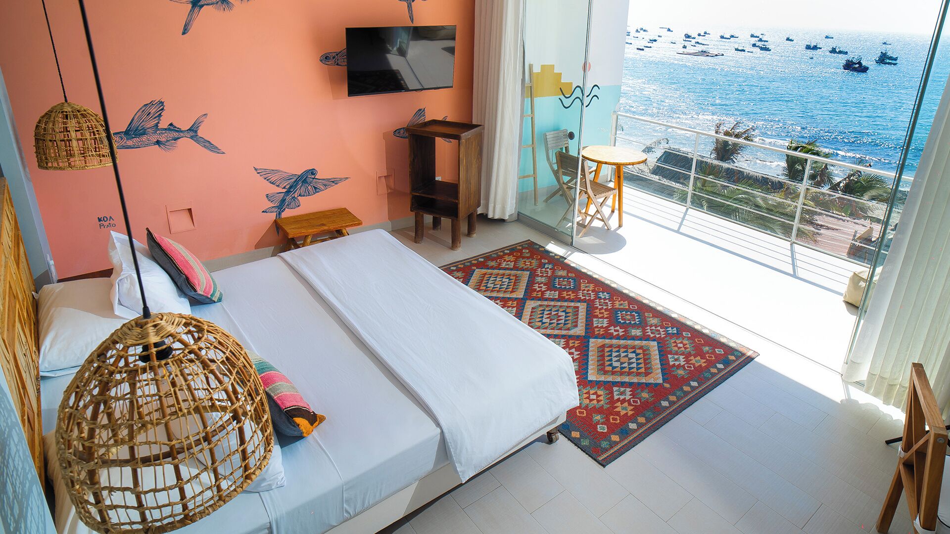 A bedroom with a bed, a TV, a small table, a chair, a hanging light, and a view of the sea.