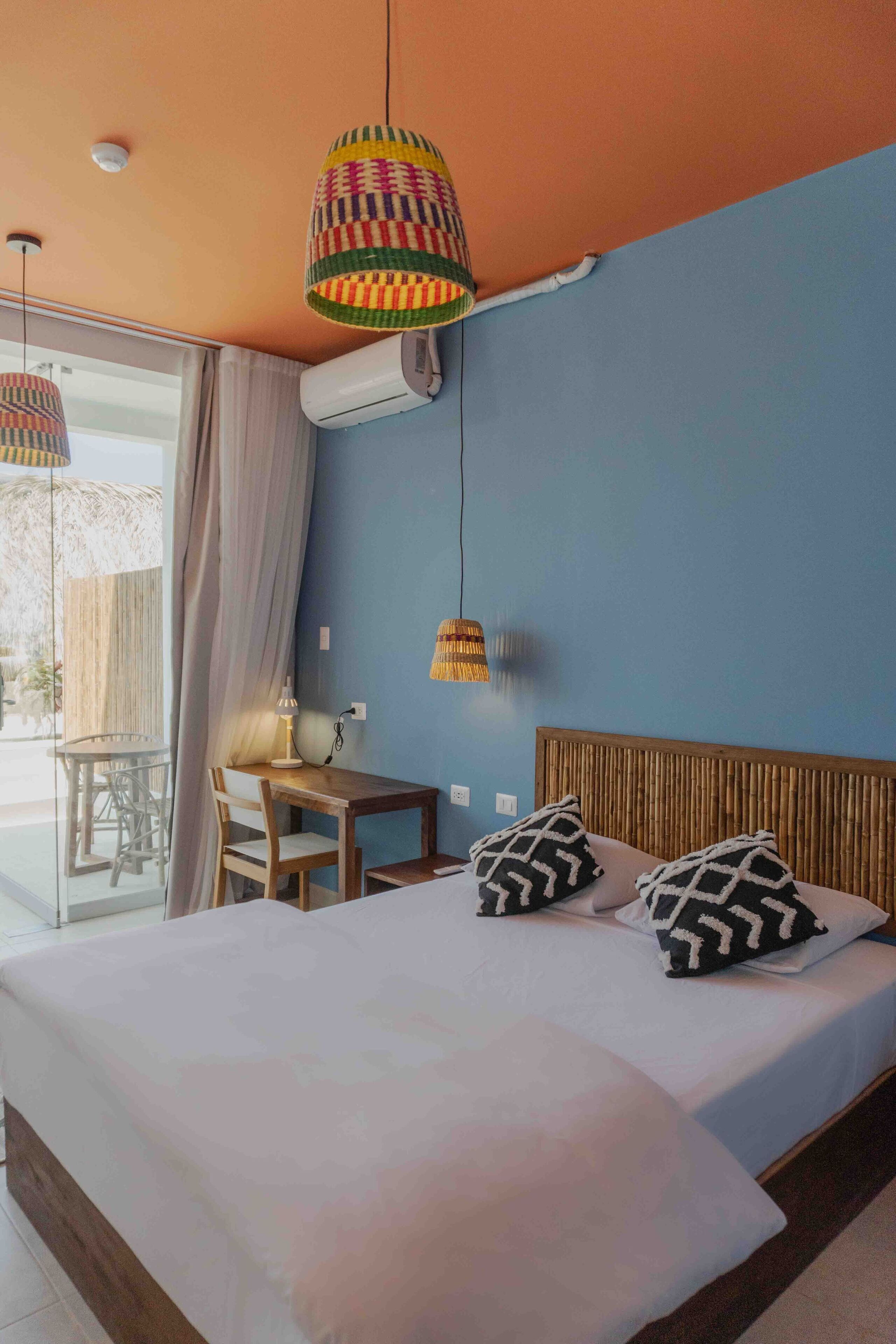 A bedroom with a bed, two patterned pillows, a wooden headboard, a table with chairs, and a hanging lamp.