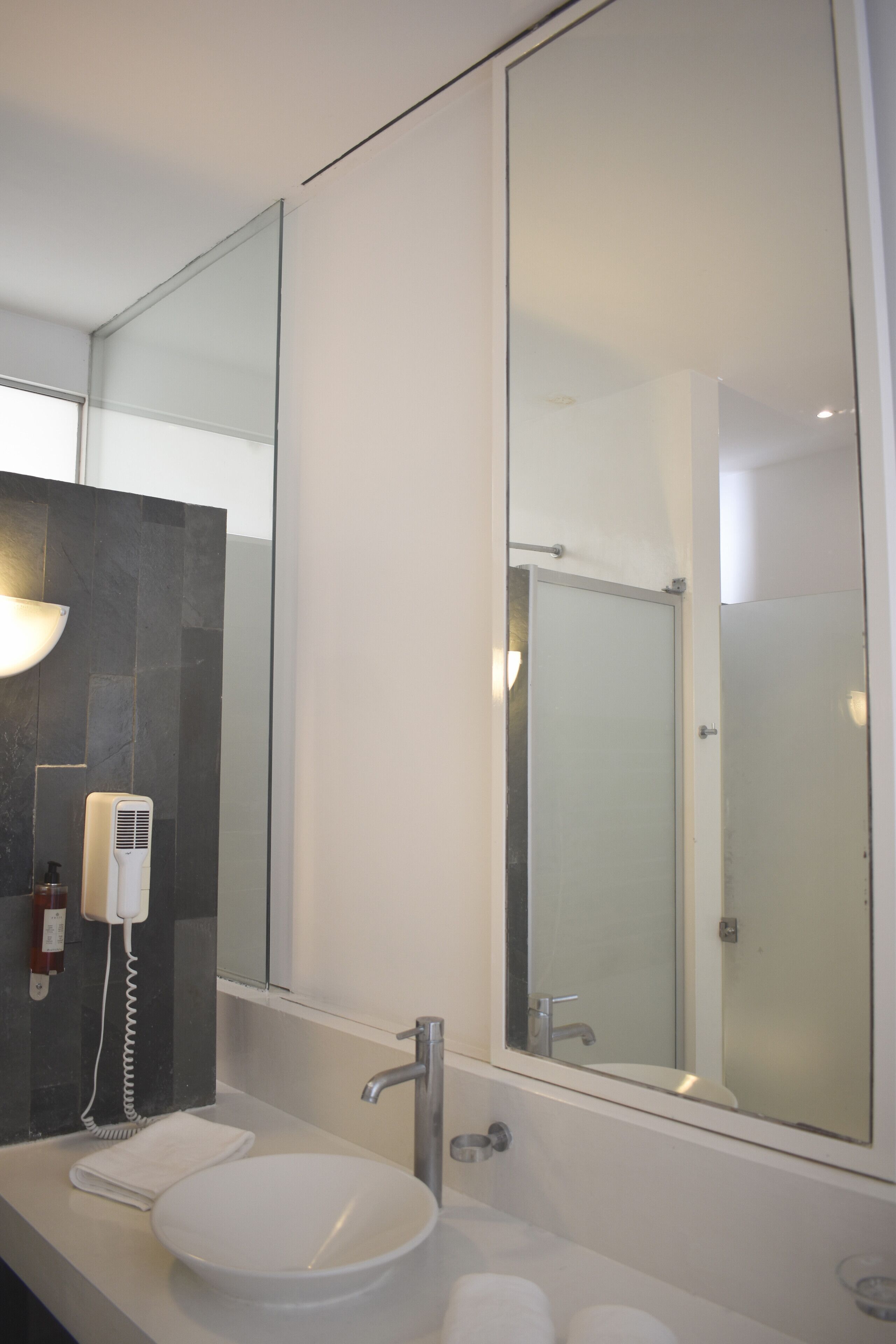 A modern bathroom with a large mirror, a white sink, a handheld hair dryer, and a glass shower enclosure.