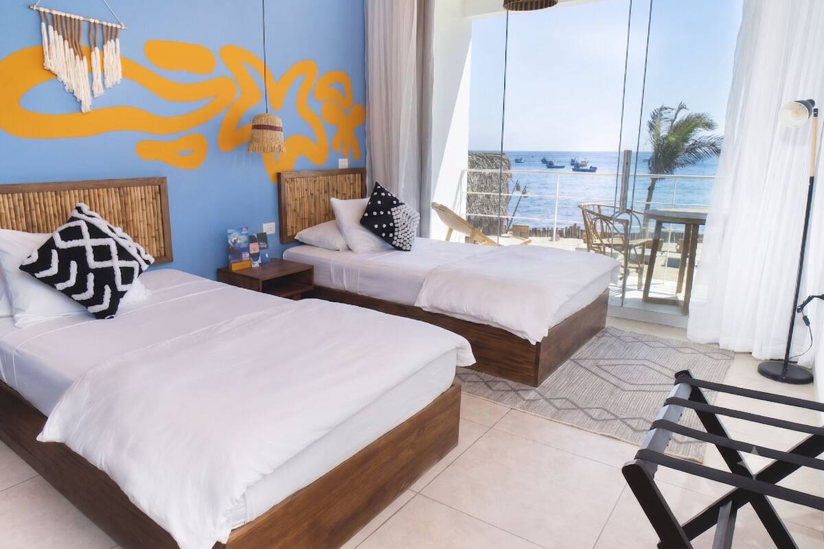 A hotel room with two beds, a large window with a view of the sea, a hanging macrame decoration, and a wooden headboard.