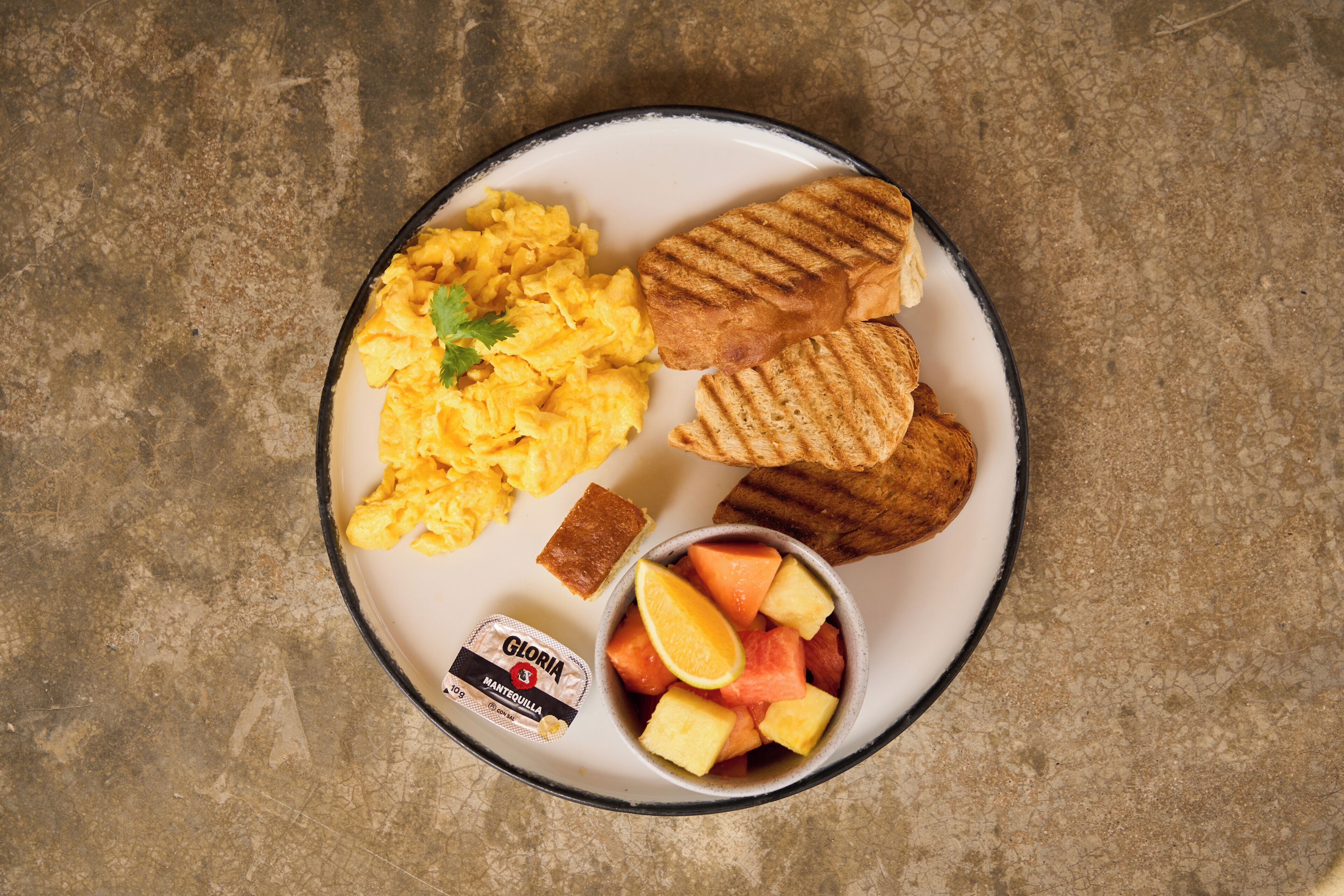 Un plato con huevos revueltos, pan a la parrilla, un trozo de pan de maíz y un bol de fruta.
