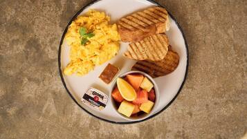 Un plato con huevos revueltos, pan a la parrilla, un trozo de pan de maíz y un bol de fruta.
