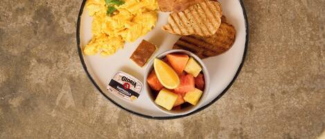 Un plato con huevos revueltos, pan a la parrilla, un trozo de pan de maíz y un bol de fruta.