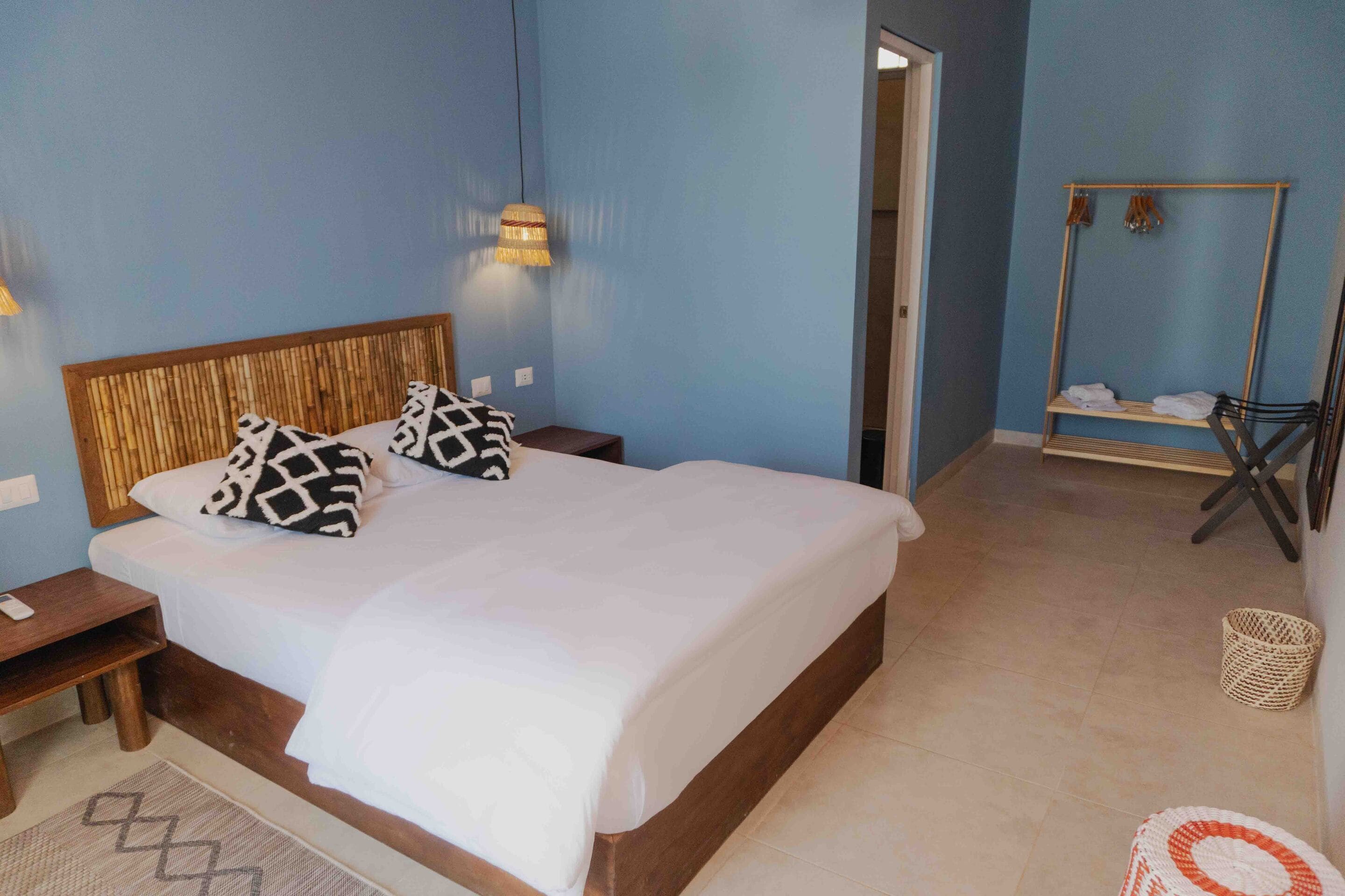 A bedroom with a wooden headboard, two bedside tables, a bed with white bedding, a hanging lamp, a wooden shelf with towels, and a woven basket on the floor.