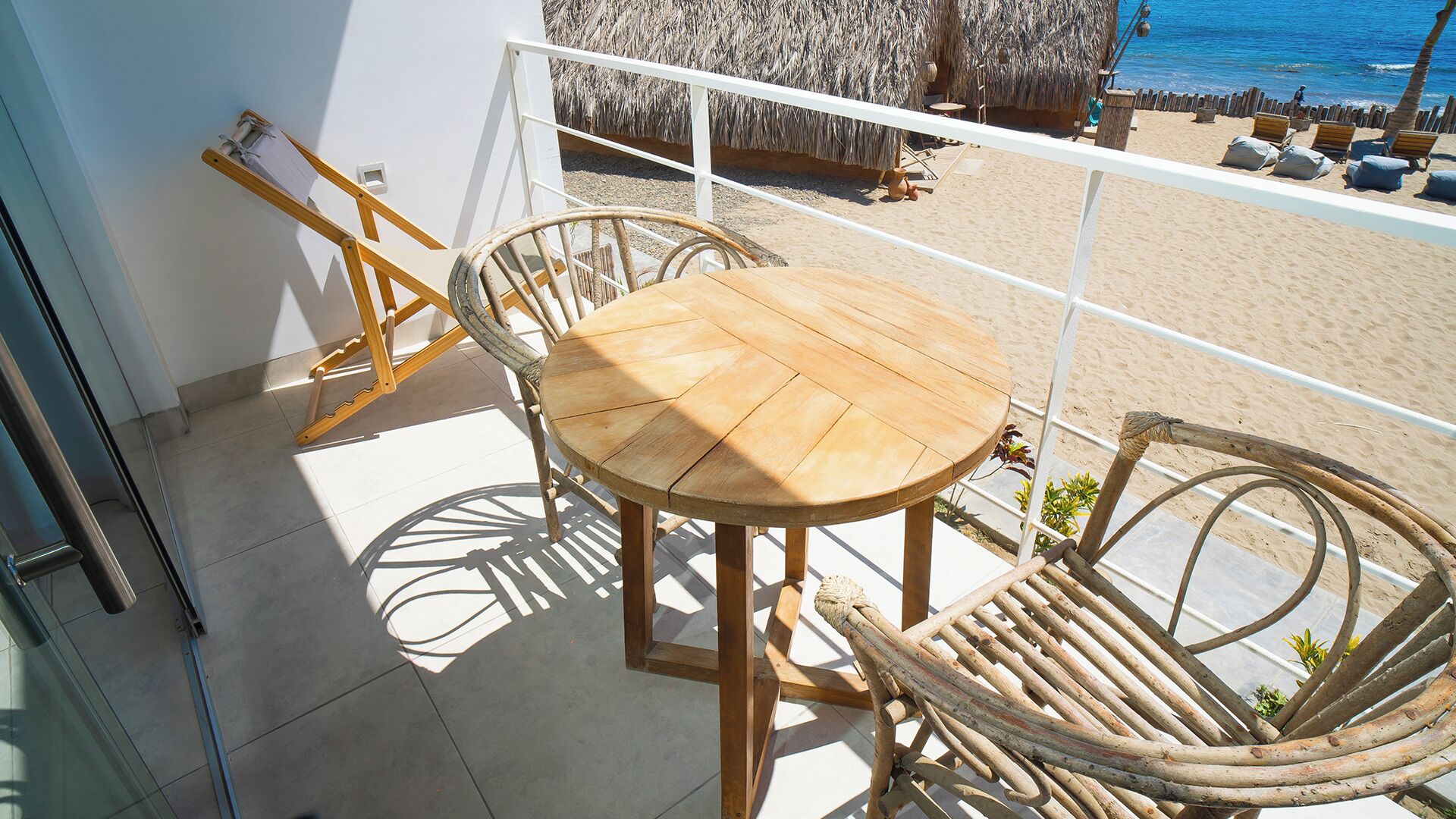 A balcony with a wooden table and wicker chairs, overlooking a beach with a thatched-roof structure.