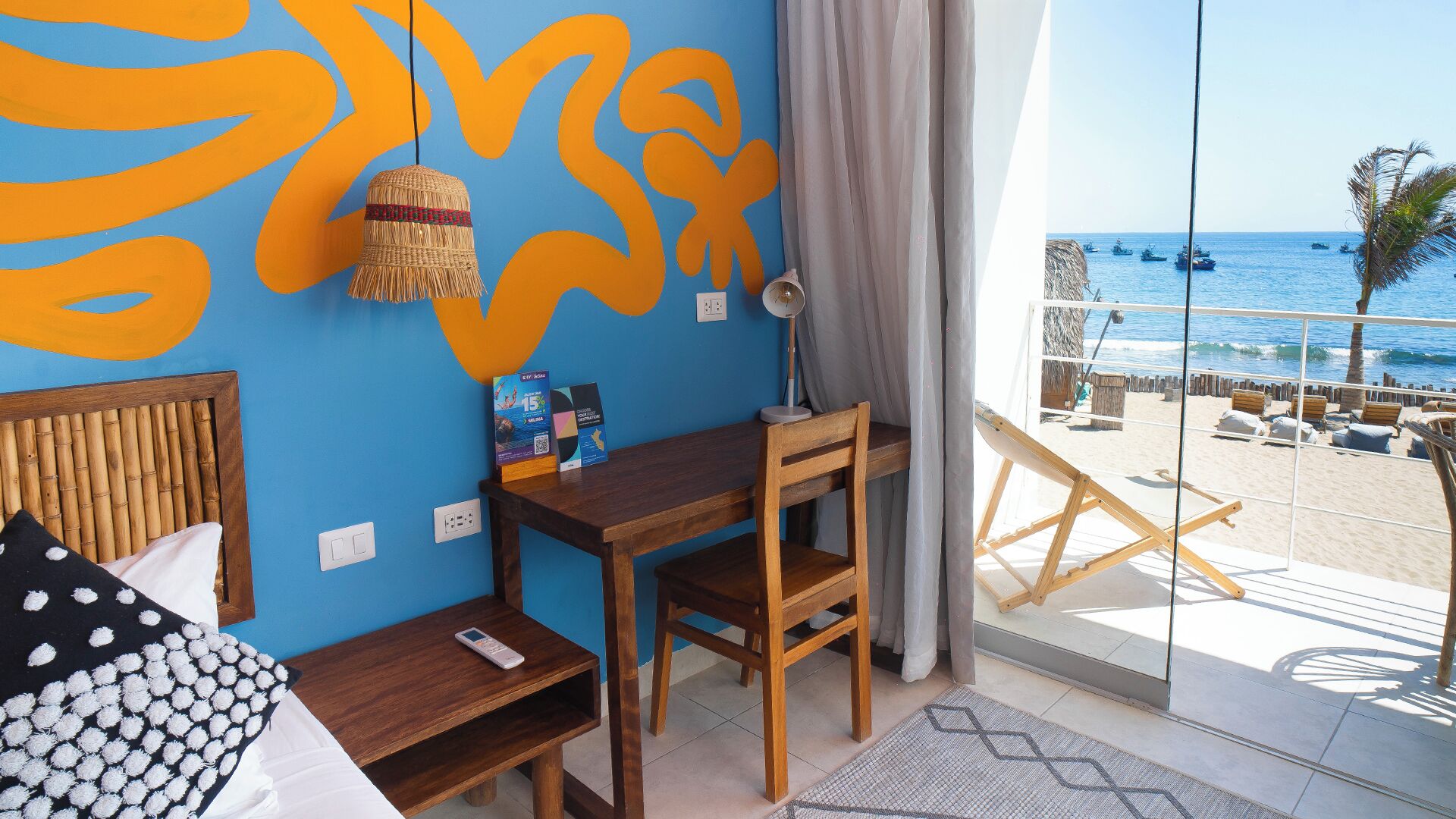 A room with a bed, desk, and chair, a large window with a view of the beach and ocean, and a wall with abstract orange and blue patterns.