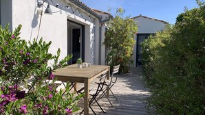 A wooden deck with a table and chairs, surrounded by greenery and a white building with a tiled roof. - Maison d'Hotes les Petites Terres (Sainte-Marie-de-Re)