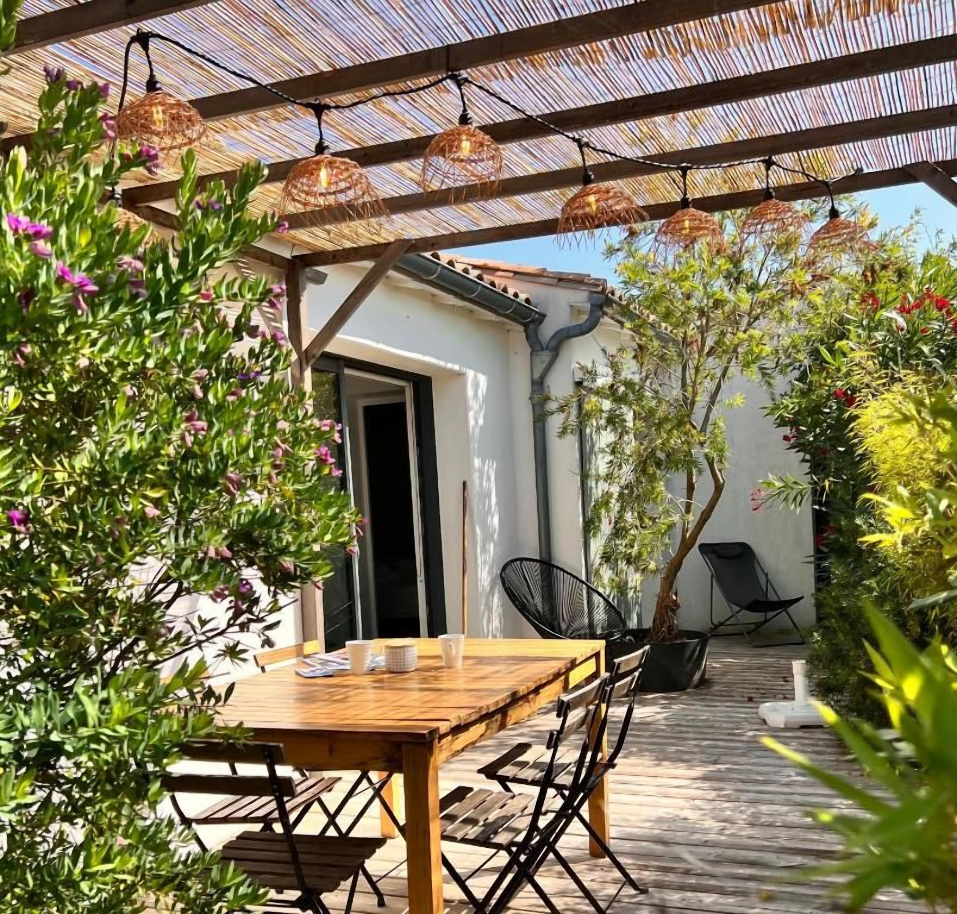 Une terrasse extérieure avec une table et des chaises en bois, une pergola agrémentée de guirlandes lumineuses et des plantes en pot.