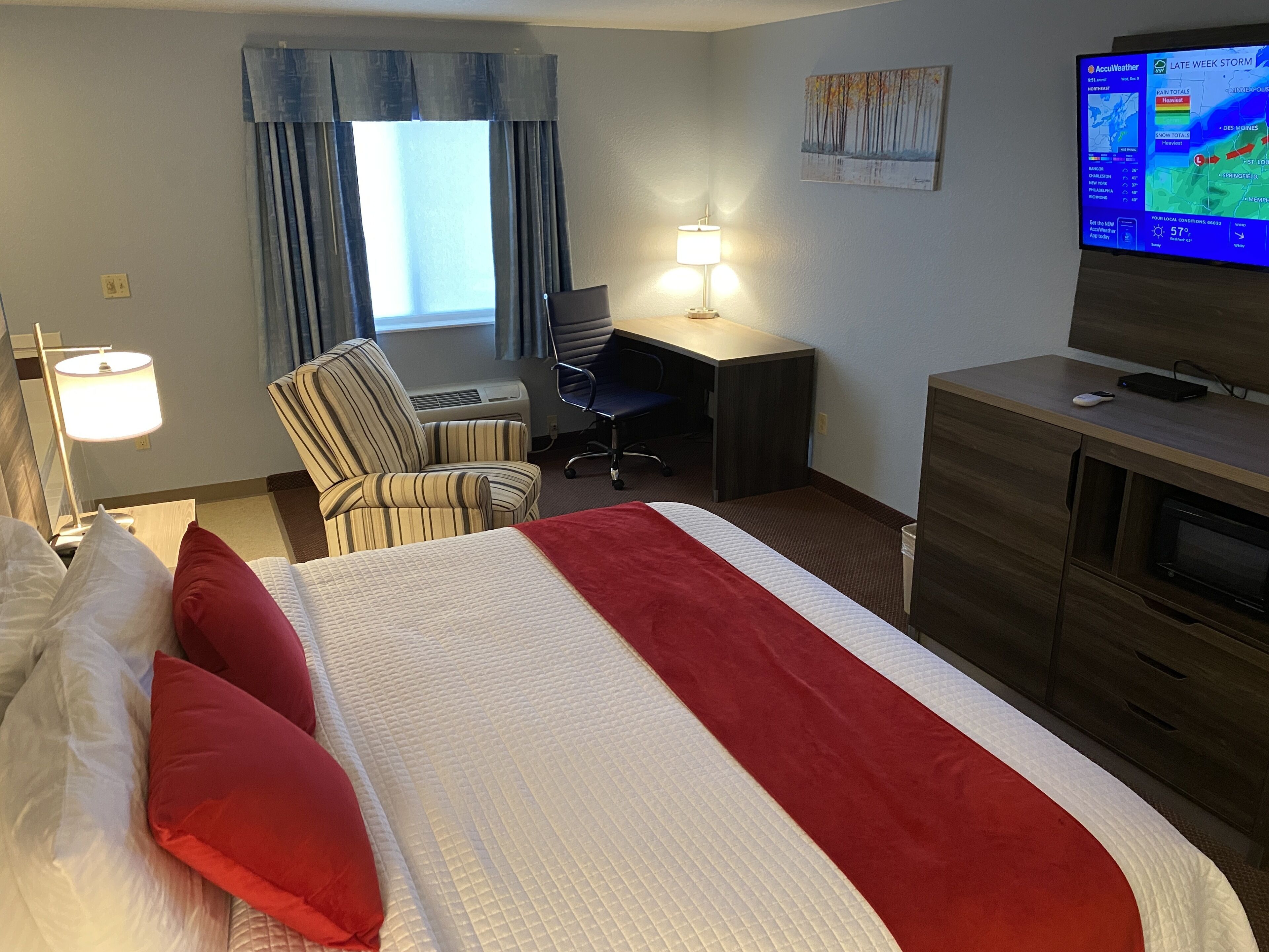 A hotel room with a bed, a desk, a chair, a lamp, a TV, and a window with curtains.
