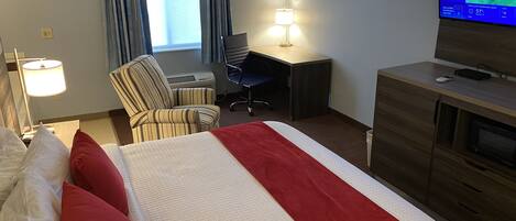A hotel room with a bed, a desk, a chair, a lamp, a TV, and a window with curtains.
